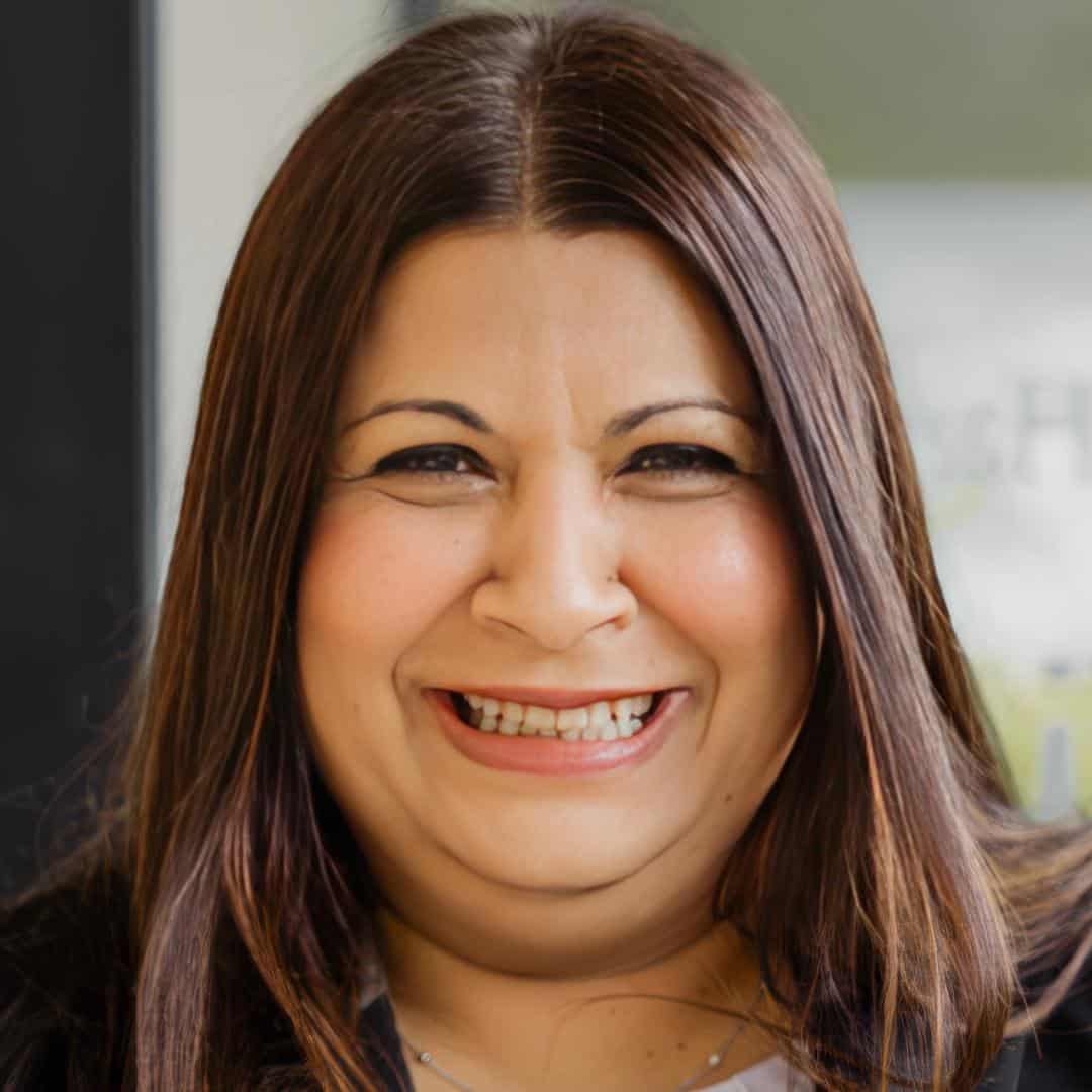 A woman with long, straight brown hair and a toothy smile looks directly at the camera. She is indoors, and part of a window is visible in the background.