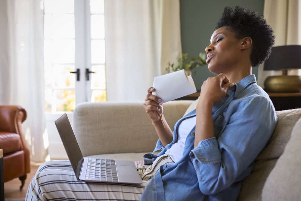A person sits on a couch with a laptop, fanning themselves with a white envelope and appearing warm.